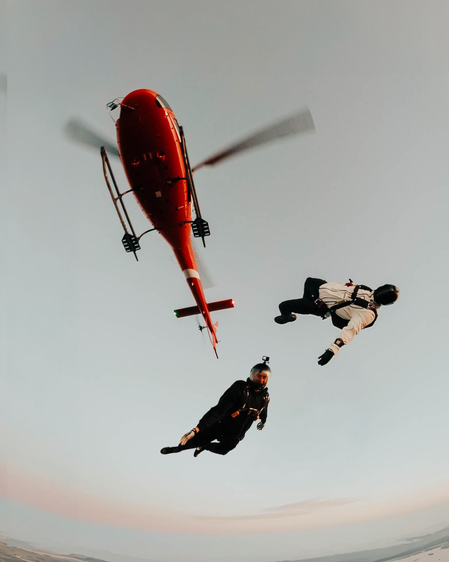 Two skydivers exiting a red helicopter above the clouds