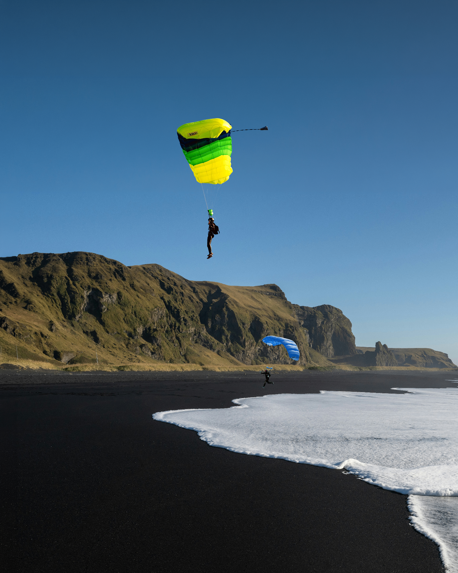 Tandem skydiver under a bright yellow and green parachute landing on Iceland's black sand beach with white surf and green cliffs